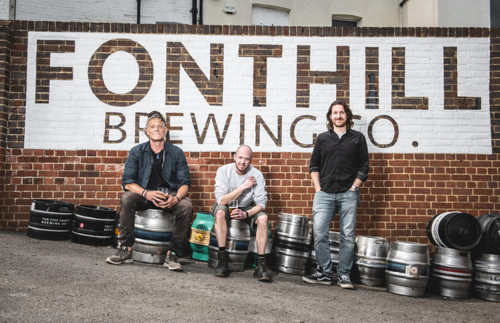 Three men, the founders of Fonthill Brewing company sitting on beer barrels at Fonthill brewery in Tunbridge Wells, with the brewery logo printed on the wall behind them